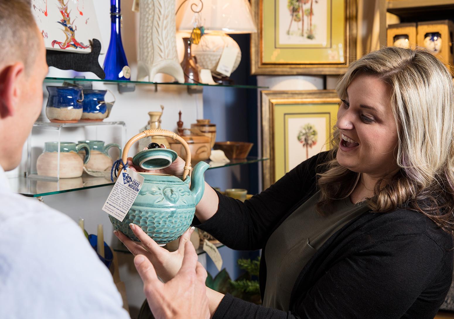 Blonde woman holds up light green ceramic tea pot on sale in the museum store