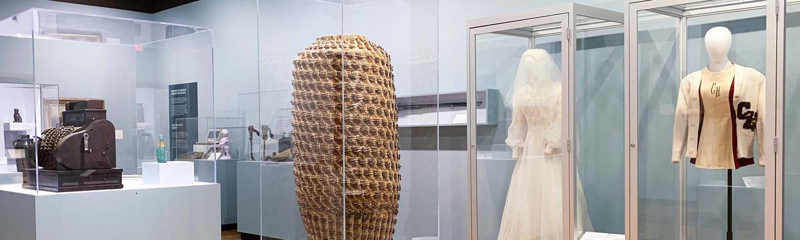 Gallery with display cases containing a large, tall narrow vase, pottery, a wedding dress and a jacket