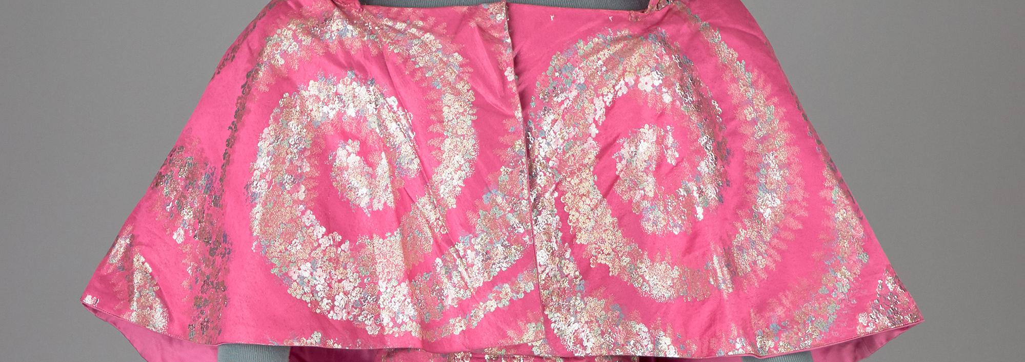 Mannequin on a gray background wearing a hot pink dress with gold and silver swirls, and a matching hooded capelet