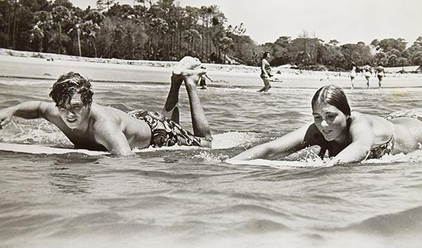 Two individuals laying down on surfboards padding in the water with a beach behind them