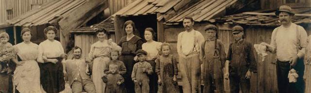 Old, sepia toned photograph depicts large group of adults and children in distressed clothing outside of a run down building