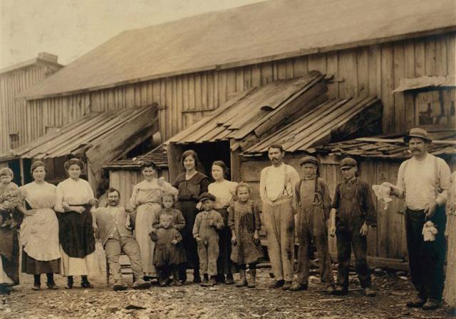 Old, sepia toned photograph depicts large group of adults and children in distressed clothing outside of a run down building