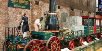 Full size train replica of The Best Friend of Charleston at the State Museum. Shows green train with red wheels and black smoke stack.