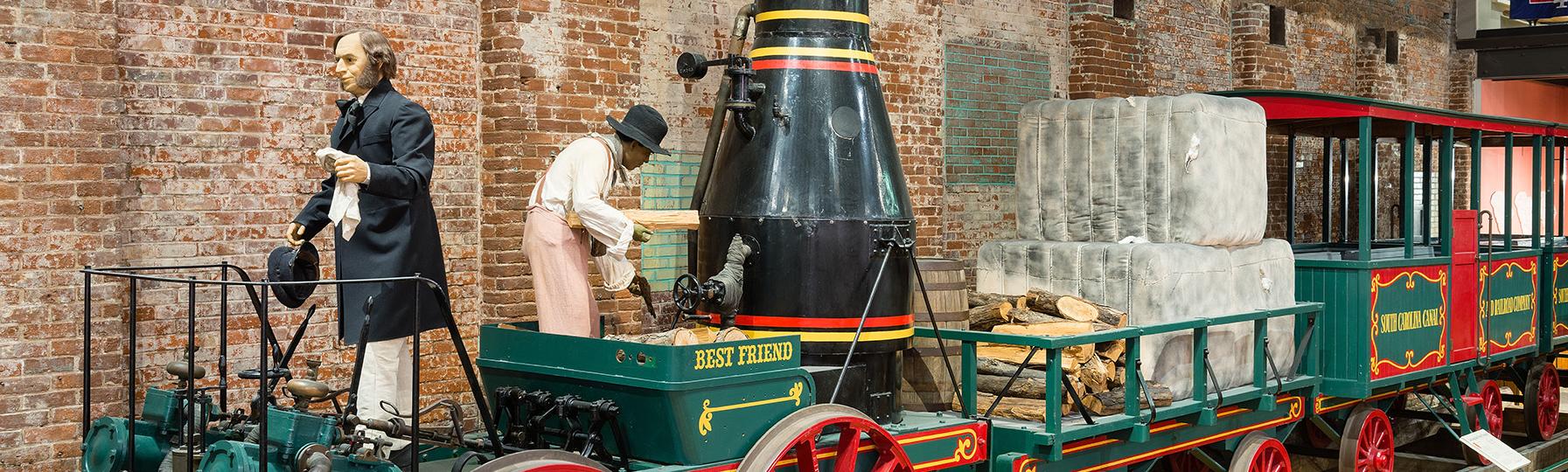 Full size train replica of The Best Friend of Charleston at the State Museum. Shows green train with red wheels and black smoke stack. 