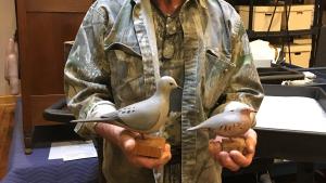 Man with mustache wearing a camouflage shirt and overalls stands holding two carved wooden birds that are painted gray with white stomachs a small black dots near their tails.