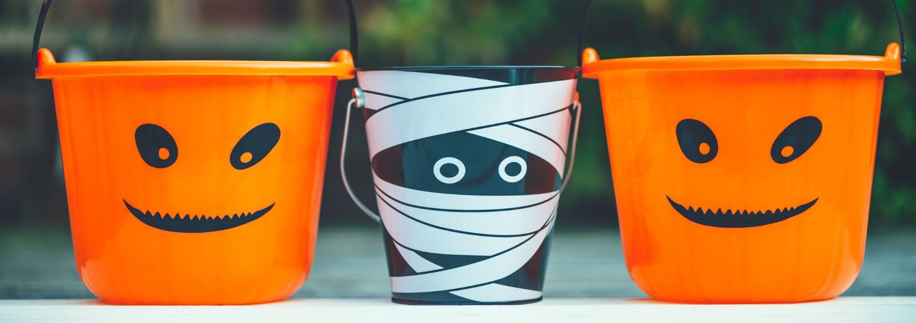 Three plastic trick or treating plastic baskets with handles. Baskets on the right and left are orange with black eyes and mouth drawn on. The center basket looks like it is wrapped in white bandages with 2 eyes peeping out from the between them.