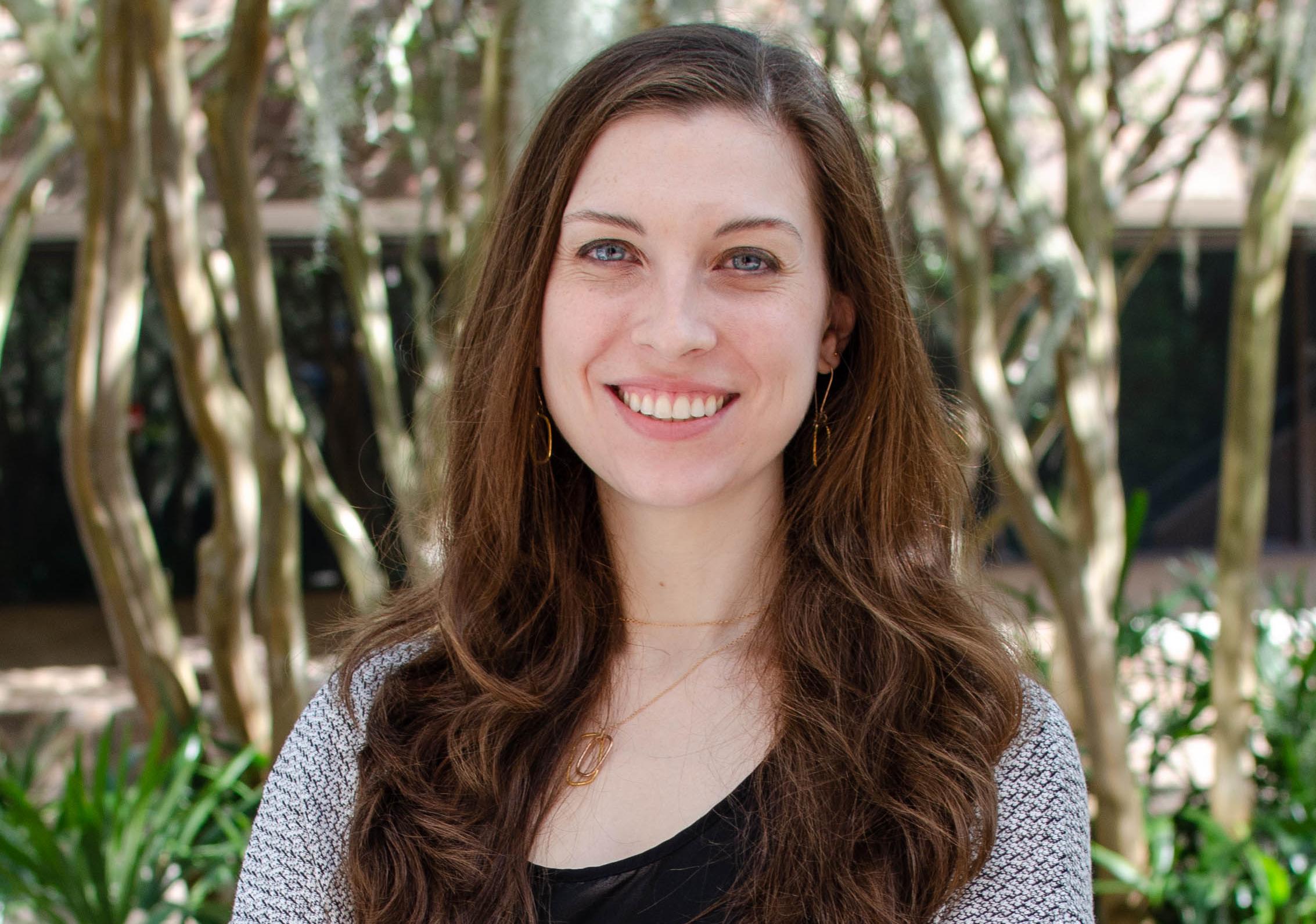Woman with long dark hair stands outside with trees in background. She is wearing a gray lightweight sweater and a dark shirt.