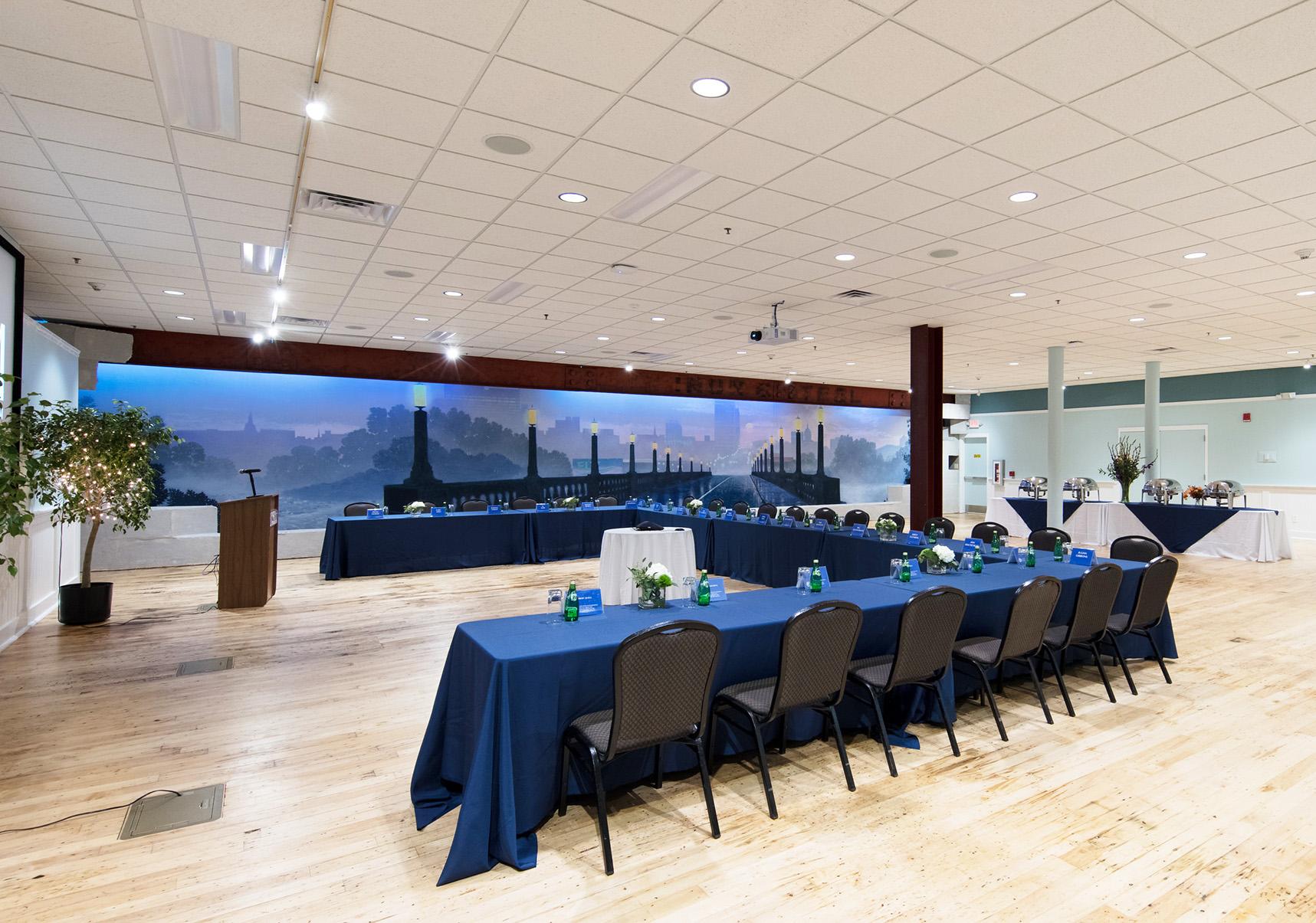Large room with light colored wooden floor has table set up in u shape facing a large screen on the wall with mural of bridge in the background