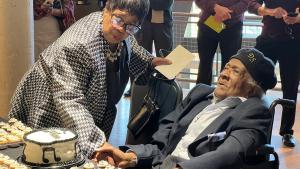 Older woman in black and white herringbone jacket leans down to assist a older man in wheelchair wearing a navy blue hat with a birthday cake on the table in front of them