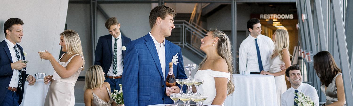 Bride and groom stand behind small, tall table with white tablecloth and members of their wedding party socializing in the background with the side of the planetarium dome exterior visible on the left side of the frame.