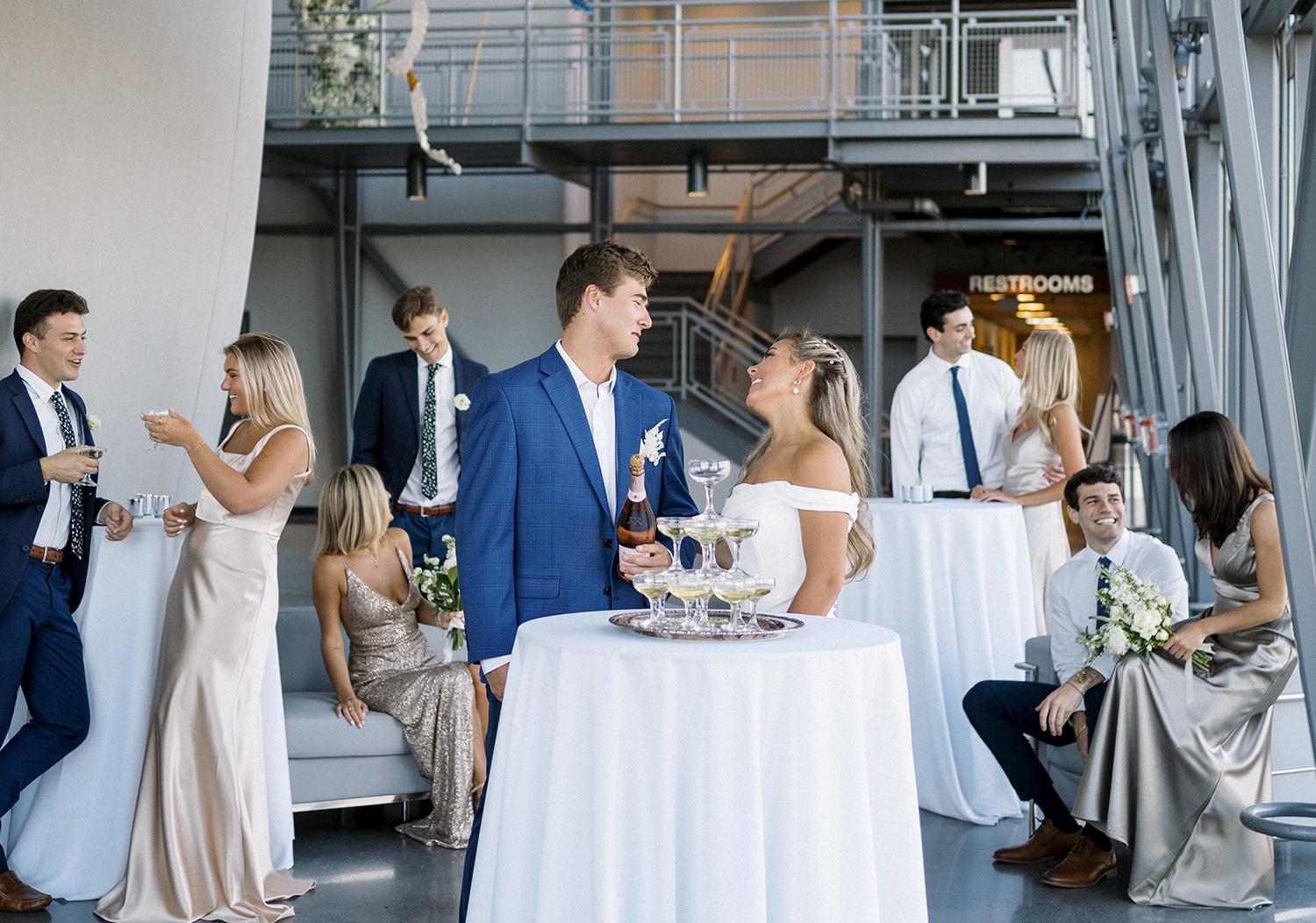 Bride and groom stand behind small, tall table with white tablecloth and members of their wedding party socializing in the background with the side of the planetarium dome exterior visible on the left side of the frame.