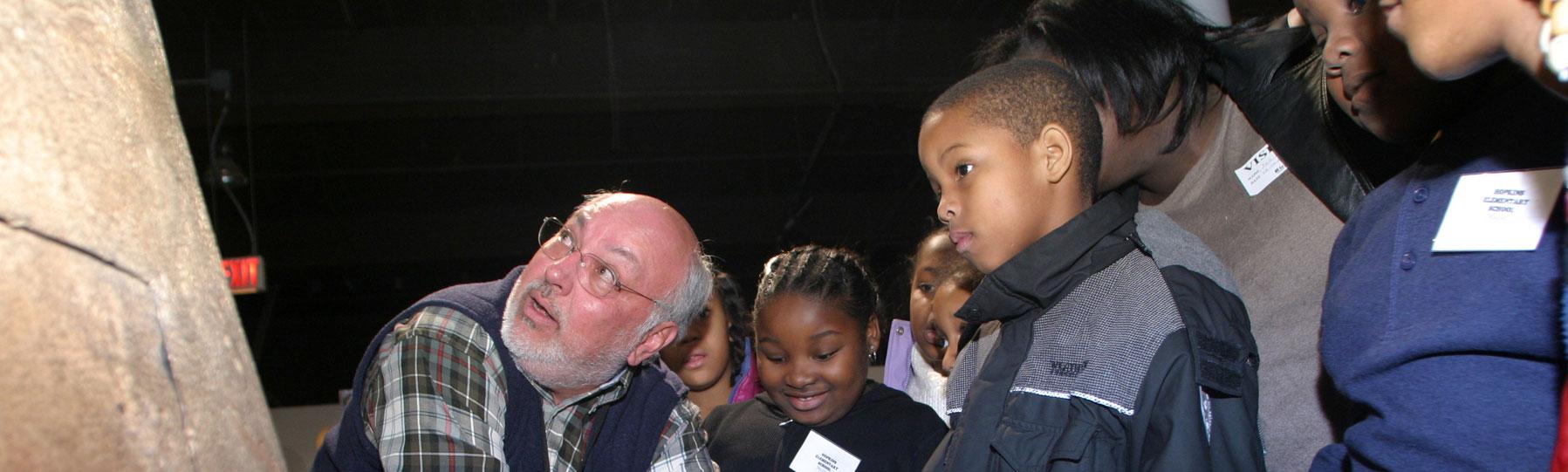 An older gentleman wearing a SCSM volunteer demonstrates an historic pot to a group of third grade students.
