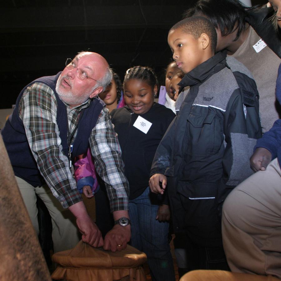 An older gentleman wearing a SCSM volunteer demonstrates an historic pot to a group of third grade students.