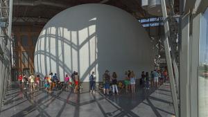 Large space with glass walls, a polished gray floor and a large gray dome structure with a line people standing in front of it