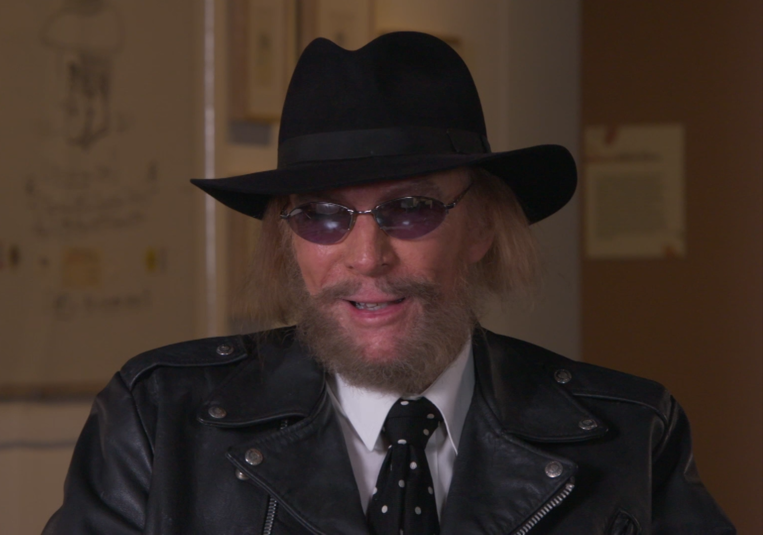 Man in black hat with dark beard and mustache wearing sunglasses and black leather jacket