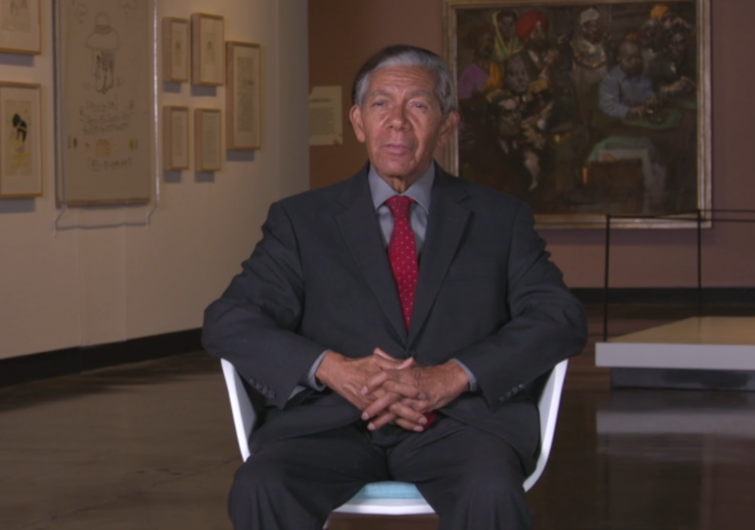 Man in gray suit and red tie sits in chair with artwork behind him