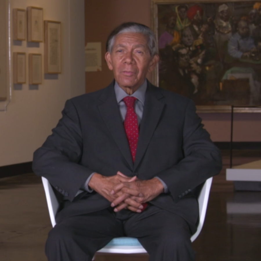 Man in gray suit and red tie sits in chair with artwork behind him