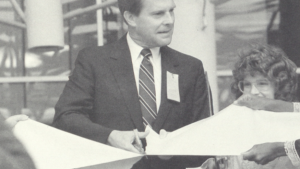 BLack and white photo of a man in a suit cutting a ribbon made of a light colored fabric