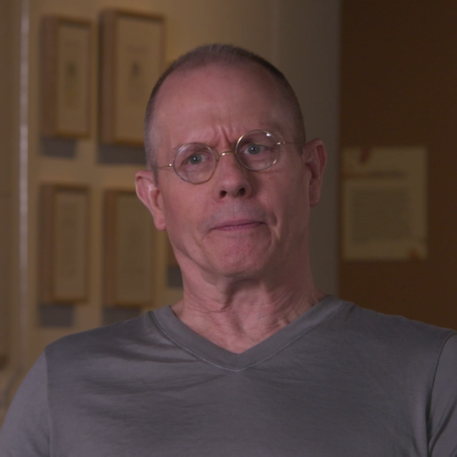 Man is gray Tshirt wearing glasses sits in chair with artwork behind him