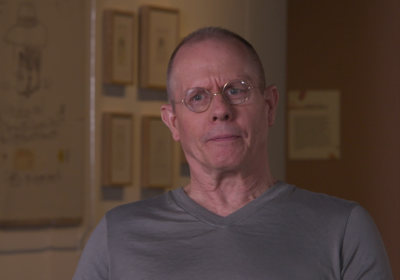 Man is gray Tshirt wearing glasses sits in chair with artwork behind him