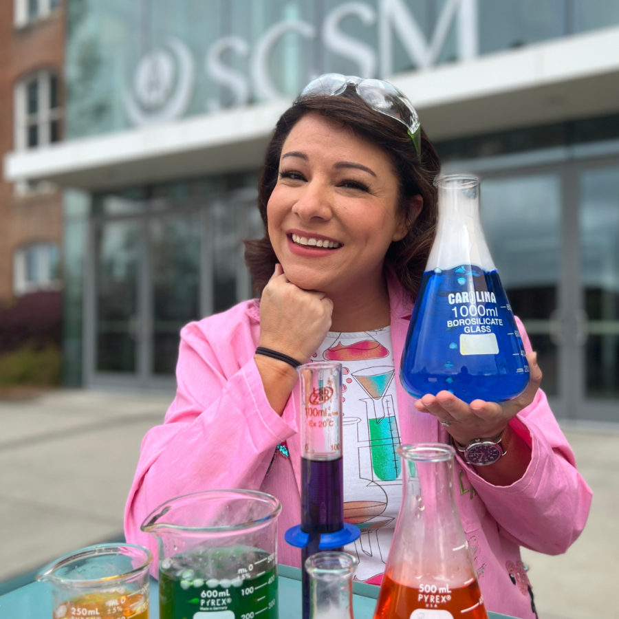 Woman with brown hair wearing a pink lab coat and holding an erlenmeyer flask with blue liquid inside smiles at camera