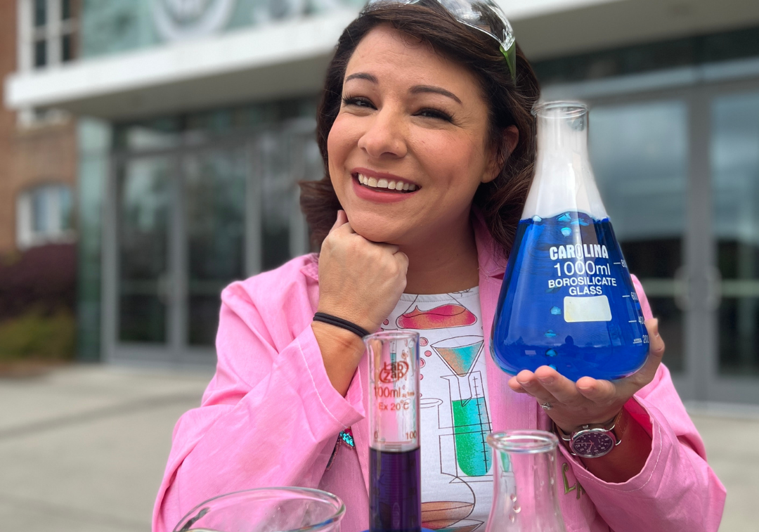Woman with brown hair wearing a pink lab coat and holding an erlenmeyer flask with blue liquid inside smiles at camera