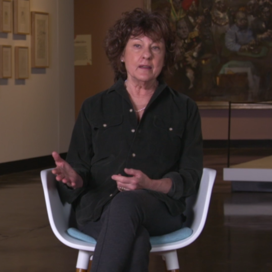 Woman in dark button-down shirt sits in chair with artwork behind her