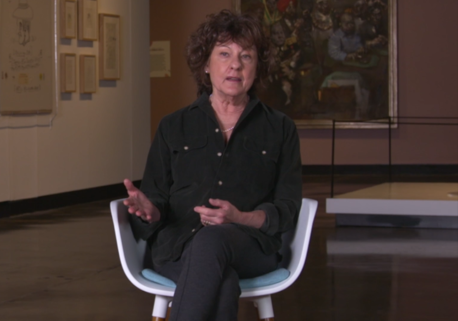 Woman in dark button-down shirt sits in chair with artwork behind her