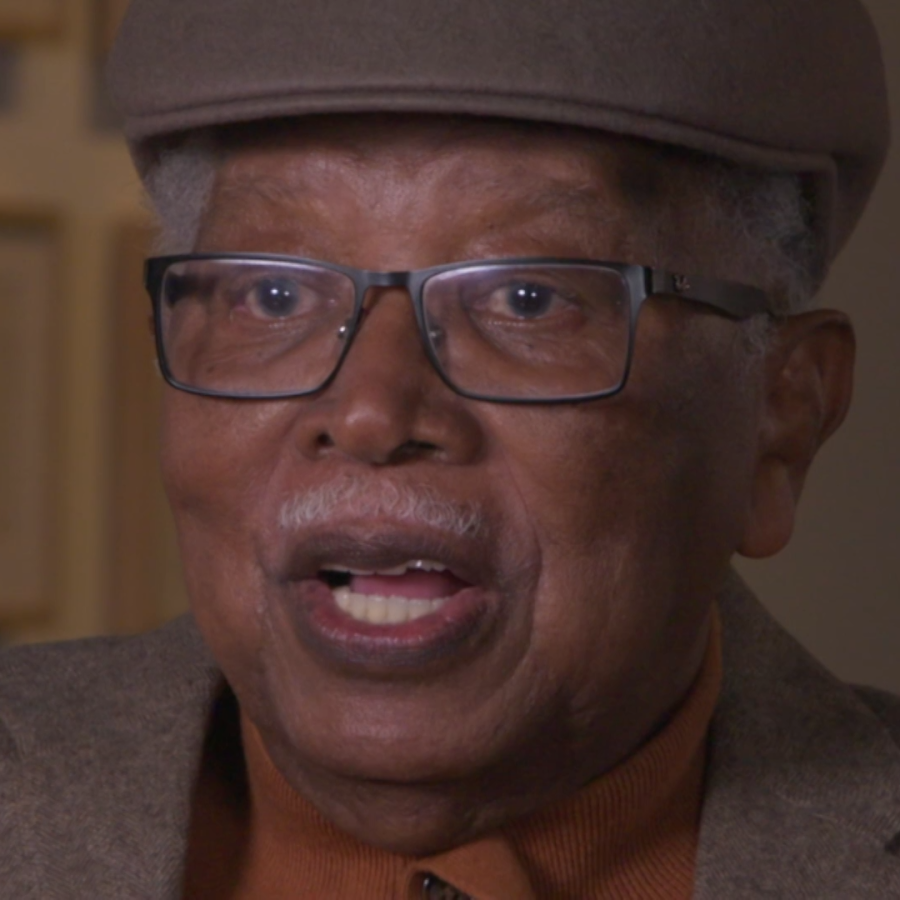 Man in glasses wearing a light brown cap faces camera with artwork behind him