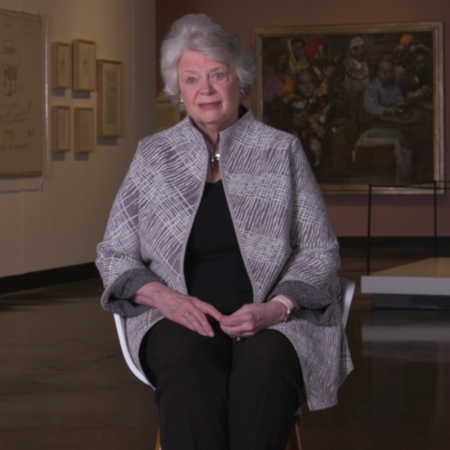 Woman in gray jacket and black top and pants sits in chair with artwork behind her.
