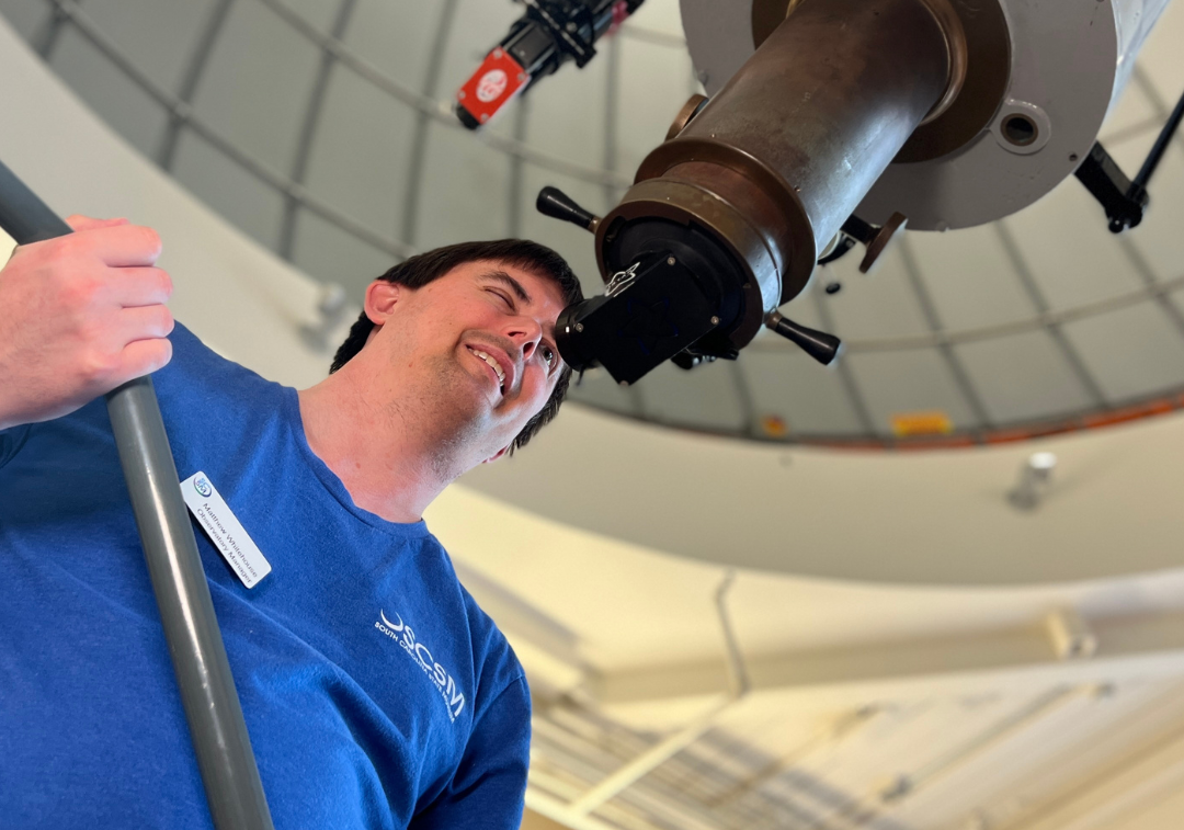 Man in long sleeved blue shirt looks through eyepiece of large white telescope