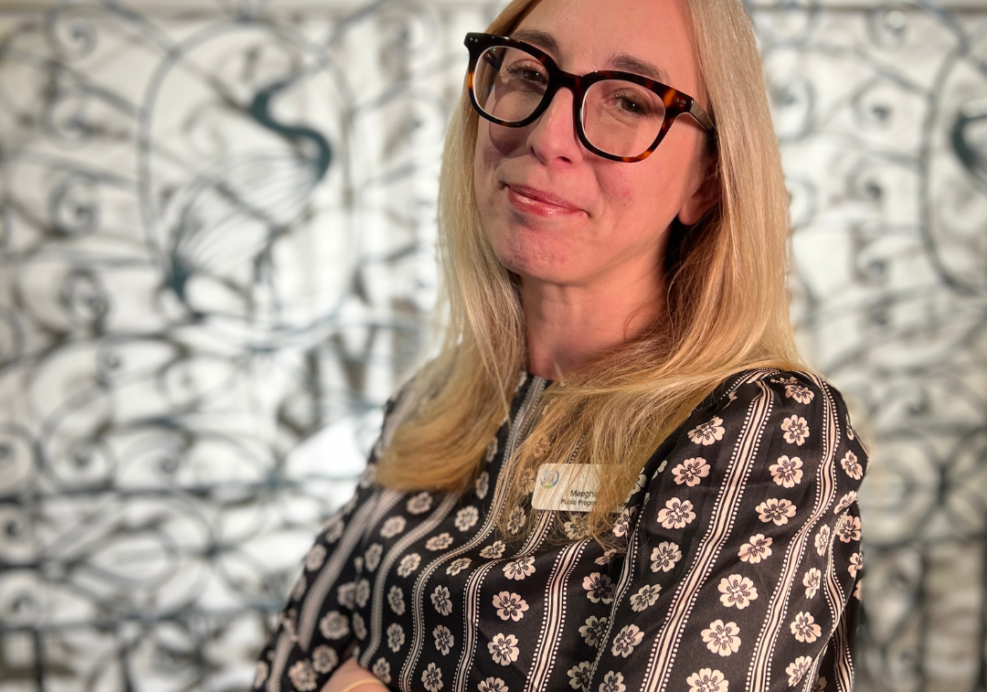 Woman with blonde hair wearing glasses stands 3/4 towards camera with arms crossed