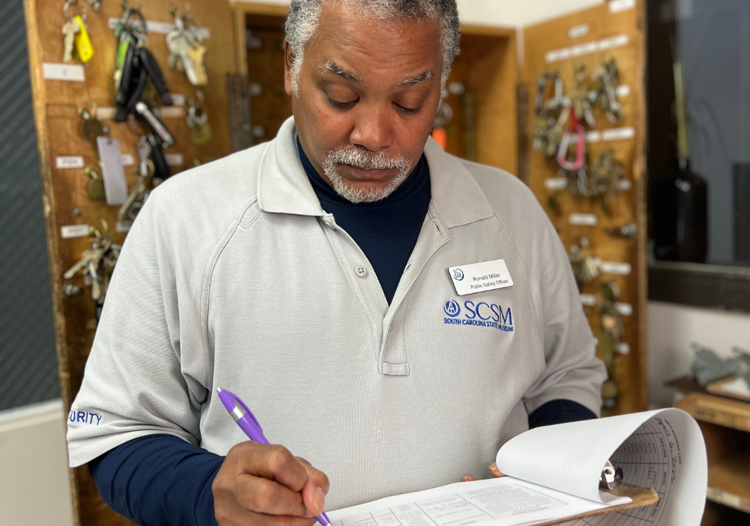Man with beard and mustache wear grey shirt with museum logo looks down at papers on a clip board