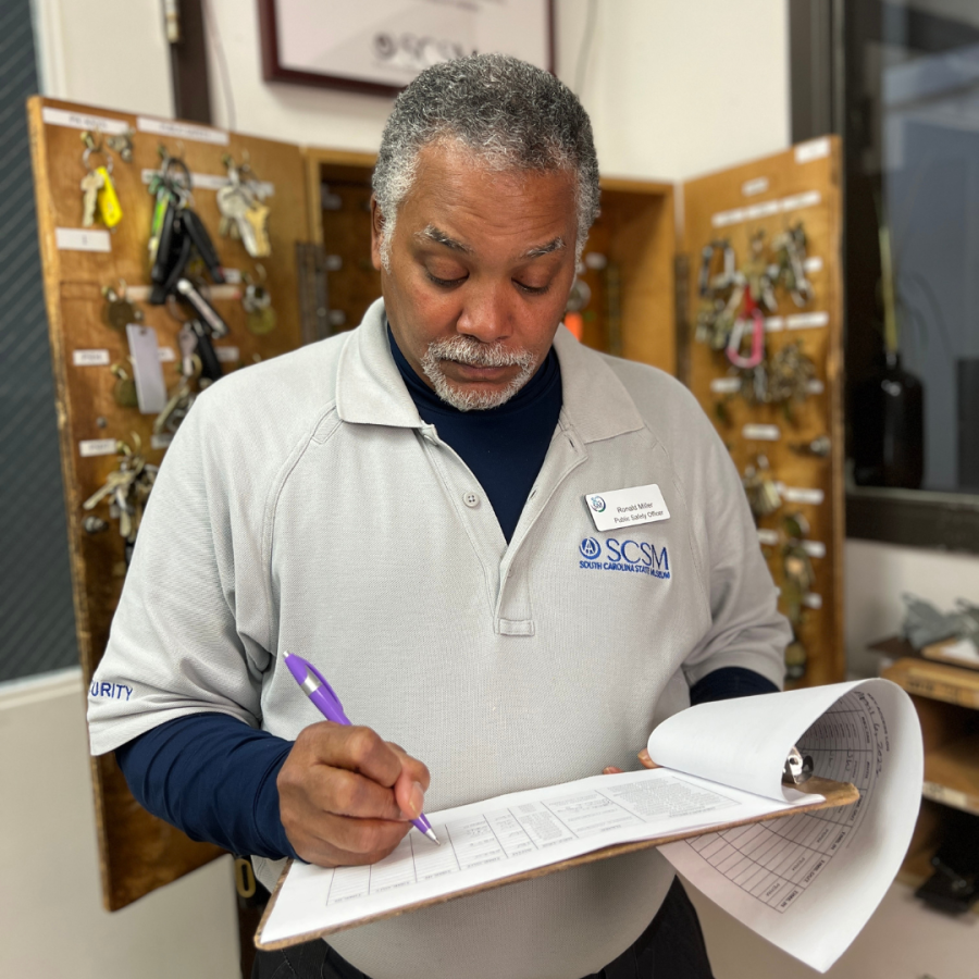 Man with beard and mustache wear grey shirt with museum logo looks down at papers on a clip board