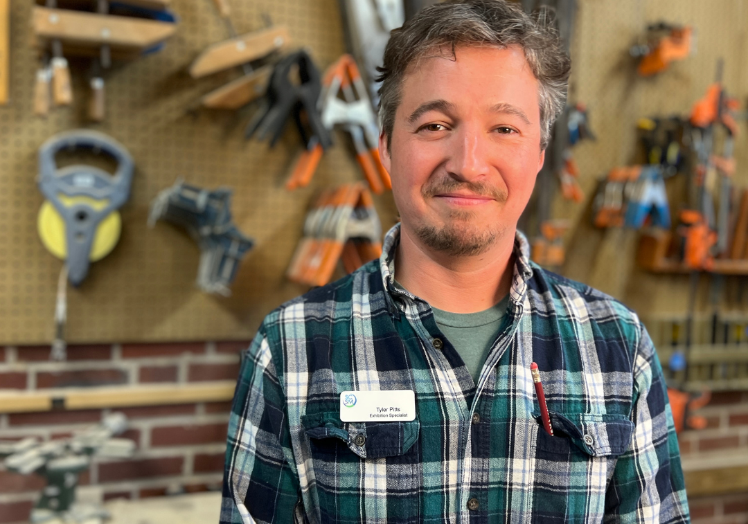Man with beard and mustache wearing green plaid shirt smiles at camera with wall of tools behind him