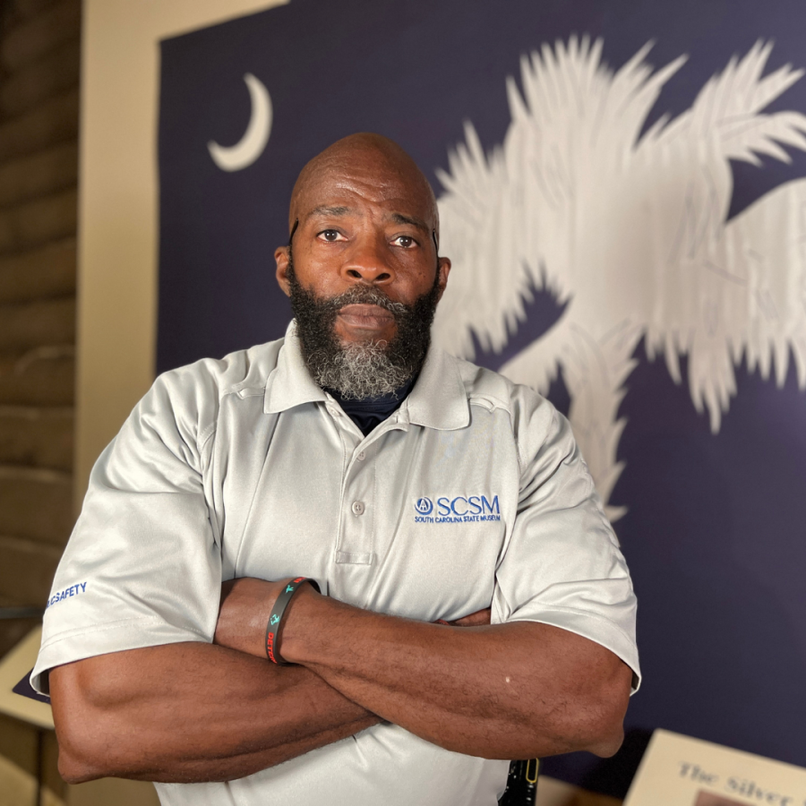 Man with beard in grey shirt with museum logo stands with arms crossed with SC state flag in the background