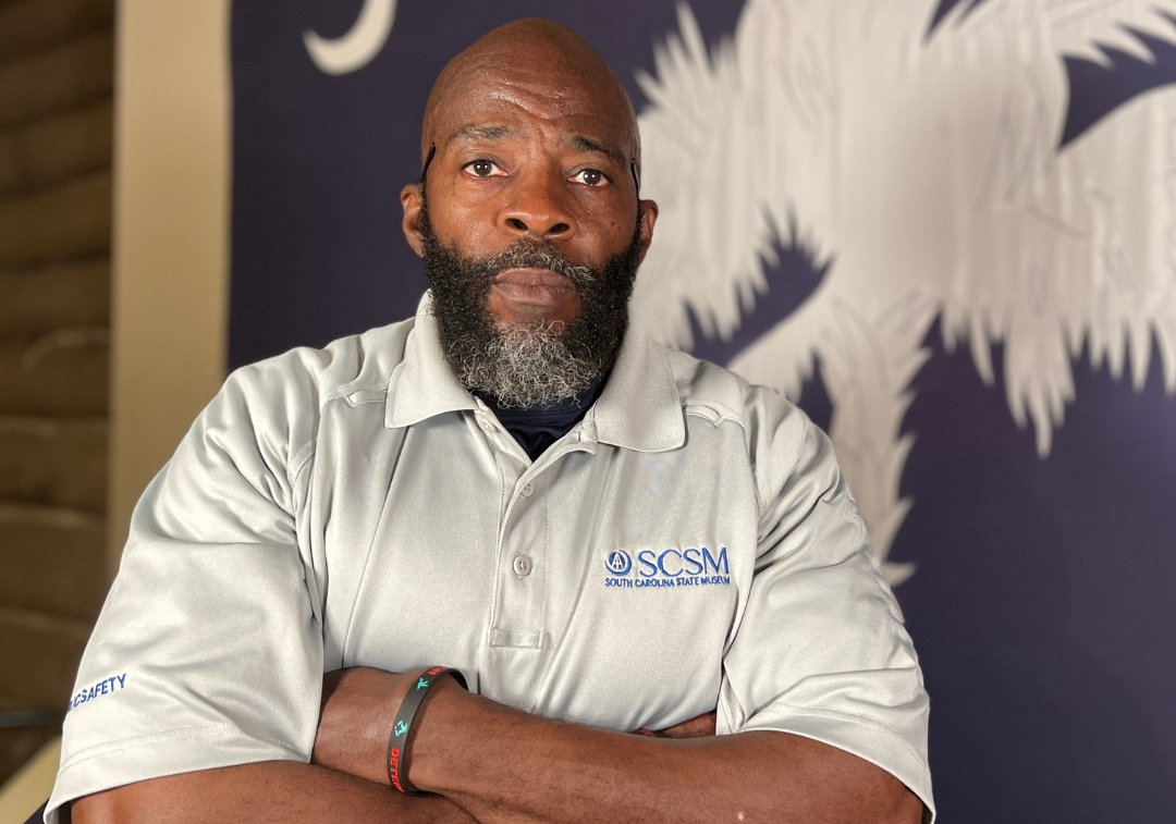 Man with beard in grey shirt with museum logo stands with arms crossed with SC state flag in the background